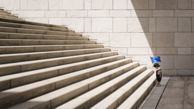 There is a small child standing in front of a huge staircase wearing a cap with a European flag.