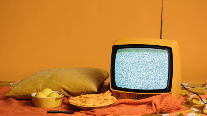 Titelbild für den Blogbeitrag. Ein Fernseher steht auf einer Orangen Decke neben einer Schale Pommes und Äpfeln.