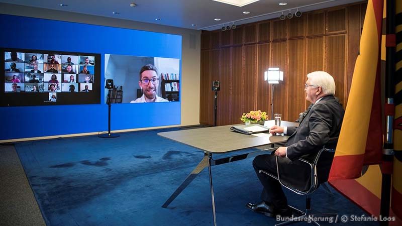 Man sieht den Bundespräsidenten, wie er in einer Liveschaltung mit vielen Teilnehmenden auf einen großen Bildschirm per Zoom-Call diskutiert. 