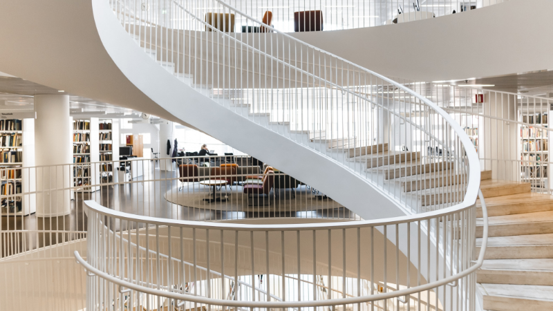 Du siehst eine Bibliothek mit einer runden Treppe die sich in die höhe schraubt. Sie steht sinnbildlich für die sich stetig weiterentwickelnden digitalen Infrastrukturen unserer Wissensgesellschaft. You see a library with a round staircase that spirals upwards. It symbolises the constantly evolving digital infrastructures of our knowledge society.