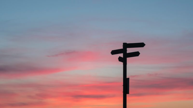 Ein Himmel bei Sonnenuntergang, auf dem rechts im Bild ein Wegweiser zu sehen ist