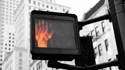 Das Foto zeigt eine Nahaufnahme eines leuchtenden roten „STOP“-Signals vor unscharfem Stadthintergrund. Das ist ein visuelles Zeichen für „Nein zu KI“ und die wachsende Resistenz gegen undemokratische Machtstrukturen in der Entwicklung Künstlicher Intelligenz.