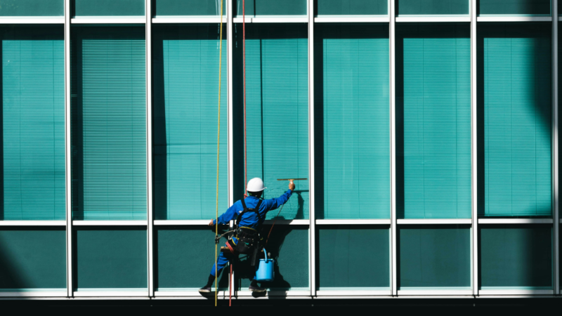 Das Foto zeigt einen Arbeiter, der eine blaue Glasfassade reinigt. Es steht symbolisch für Transparenz und das Hinterfragen von Annahmen über Desinformation.