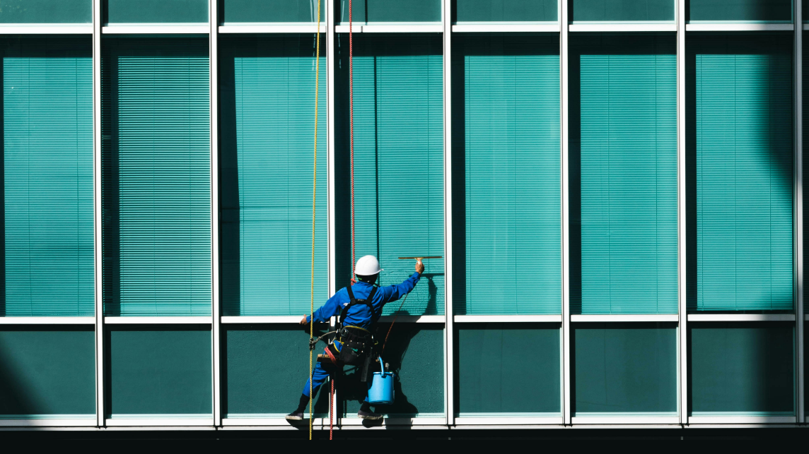 Das Foto zeigt einen Arbeiter, der eine blaue Glasfassade reinigt. Es steht symbolisch für Transparenz und das Hinterfragen von Annahmen über Desinformation.