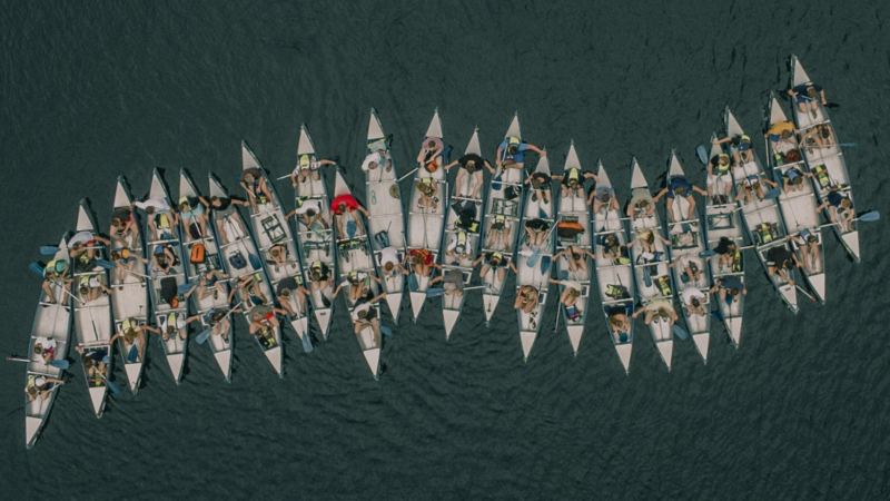 Das Foto zeigt eine Reihe von Rudernden, die sich aneinander festhalten, sodass ihre Boote eine Reihe bilden. Das ist ein Sinnbild dafür, dass es eine vereinte Strategie braucht, um Desinformationsforschende in Europa vor koordinierten Angriffen zu schützen.