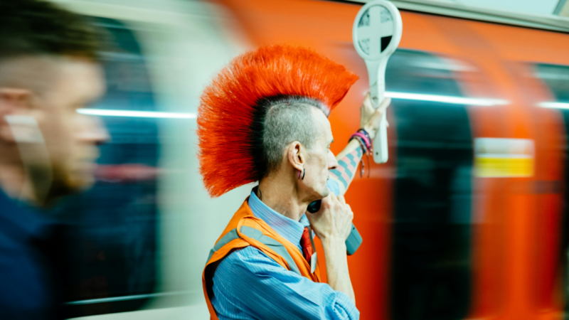 Das Foto zeigt einen Schaffner mit Warnweste und orangenem Irokesenhaarschnitt, der den Zugverkehr regelt. Er steht sinnbildlich für nationale Strategien für Plattformalternativen.