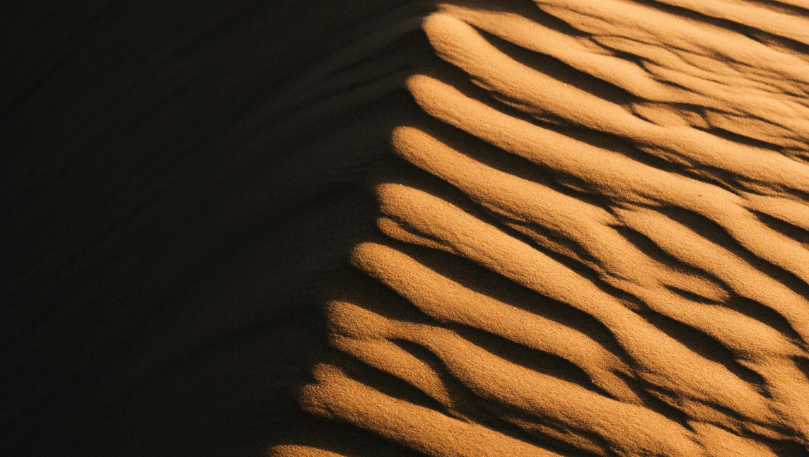 Detusch: Nahaufnahme einer Sanddüne mit wellenförmigen Mustern aus Licht und Schatten, die vom Wind geformt wurden. Englisch: Close-up of a sand dune with wave-like patterns of light and shadow formed by the wind.