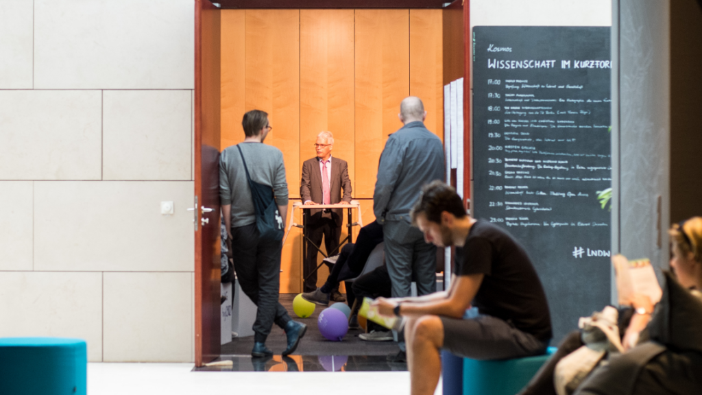 The photo shows the entrance area of an event: while people wait and an older man speaks at a standing lectern in an open space symbolising democracy without borders.