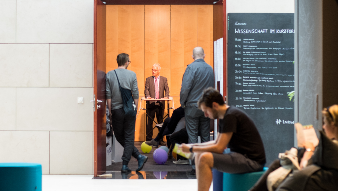 The photo shows the entrance area of an event: while people wait and an older man speaks at a standing lectern in an open space symbolising democracy without borders.