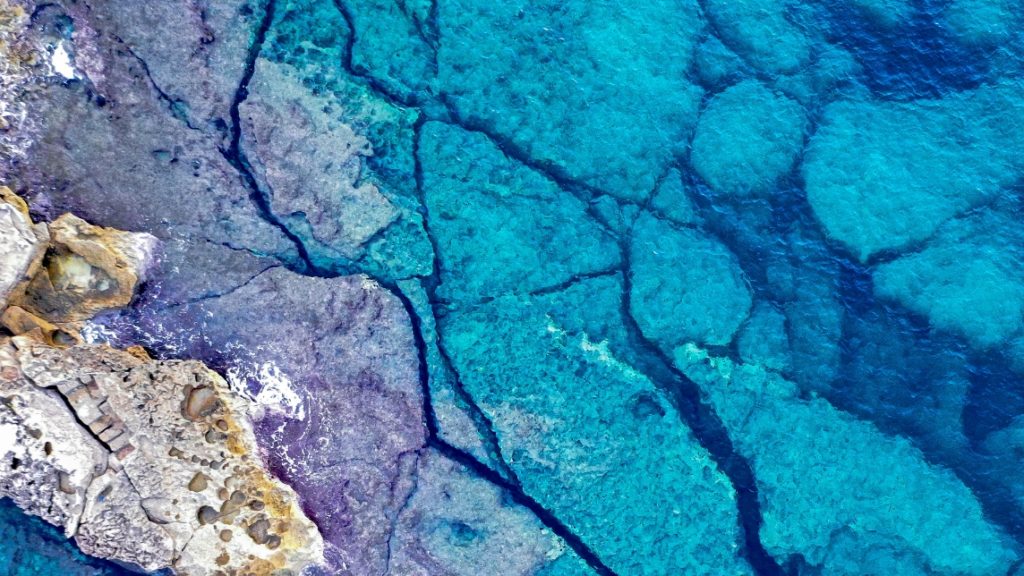 An aerial view of rocks beneath clear turquoise water symbolically represents looking beneath the surface to uncover the real impacts of AI.