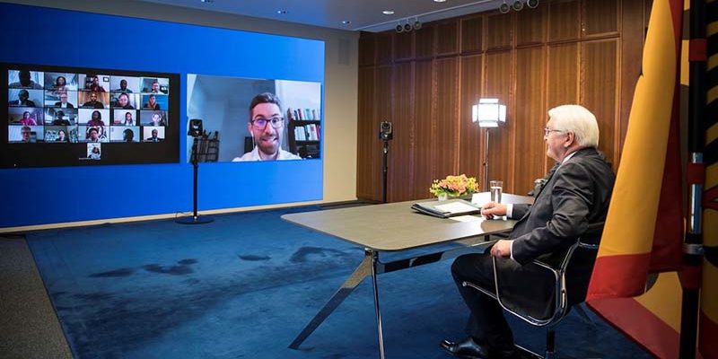 Man sieht den Bundespräsidenten, wie er in einer Liveschaltung mit vielen Teilnehmenden auf einen großen Bildschirm per Zoom-Call diskutiert.