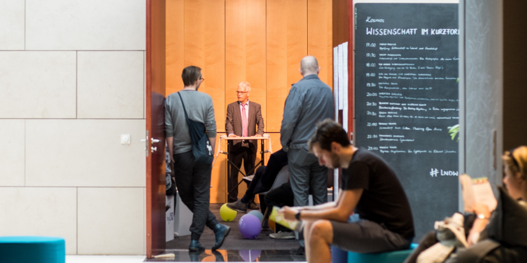 Blog-Demokratie-ohne-Grenzen-Democracy Without Borders Das Foto zeigt den Eingangsbereich einer Veranstaltung: Während die Menschen warten, hält ein älterer Mann an einem Stehpult auf einem offenen Platz eine Rede – ein Symbol für Demokratie ohne Grenzen.