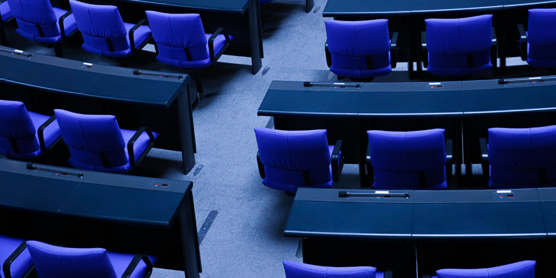 Rows of empty blue chairs in a plenary hall, symbolizing the question of whether AI can strengthen democracy.