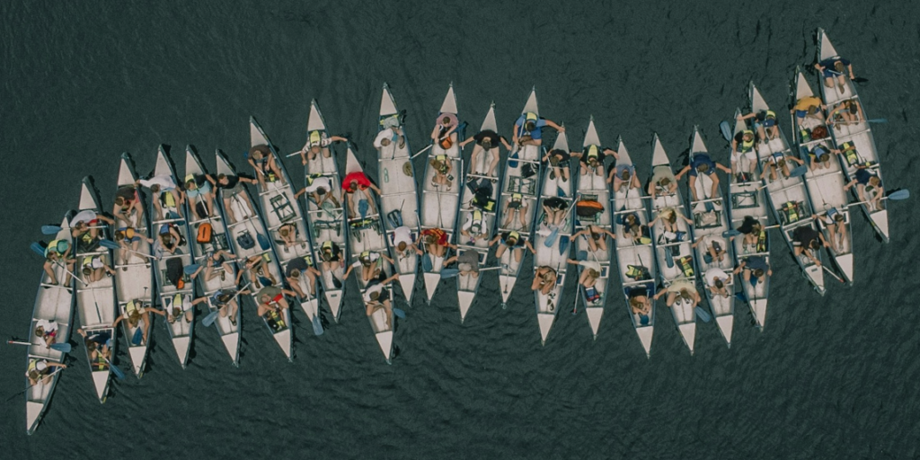 Rowers hold on to each other in boats forming a row. The image illustrates that defending Europe’s disinformation researchers against coordinated attacks needs a united strategy.