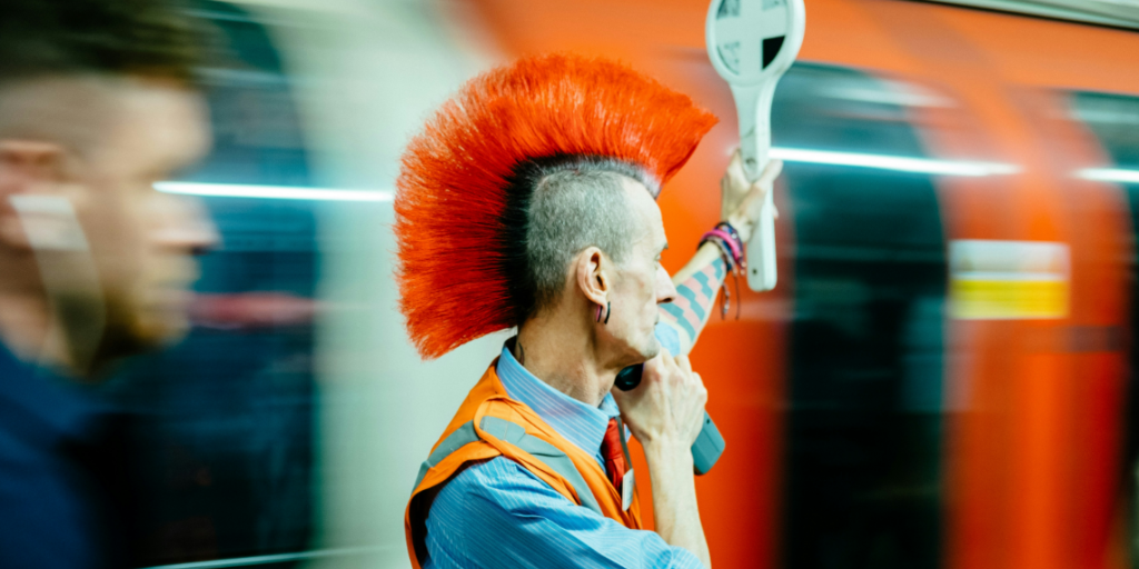 Titelbild_Platformalternatives Das Foto zeigt einen Schaffner mit Warnweste und orangenem Irokesenhaarschnitt, der den Zugverkehr regelt. Er steht sinnbildlich für nationale Strategien für Plattformalternativen.