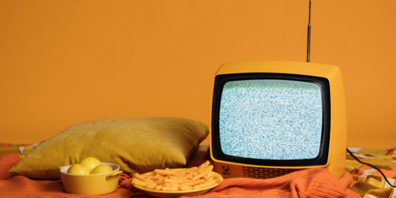 Titelbild für den Blogbeitrag. Ein Fernseher steht auf einer Orangen Decke neben einer Schale Pommes und Äpfeln.